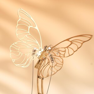 Puede incluir: Una detallada escultura de mariposa de metal dorado con intrincados dise&ntilde;os de alas. La escultura presenta elementos mec&aacute;nicos, incluyendo resortes y articulaciones, sugiriendo una est&eacute;tica cin&eacute;tica o steampunk. La mariposa se encuentra sobre un fondo c&aacute;lido y neutro.