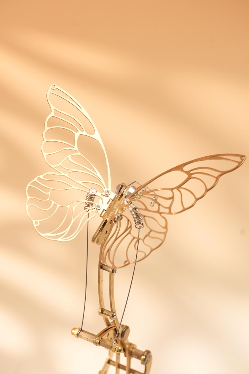 Puede incluir: Una detallada escultura de mariposa de metal dorado con intrincados dise&ntilde;os de alas. La escultura presenta elementos mec&aacute;nicos, incluyendo resortes y articulaciones, sugiriendo una est&eacute;tica cin&eacute;tica o steampunk. La mariposa se encuentra sobre un fondo c&aacute;lido y neutro.