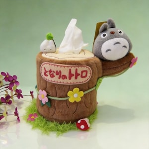 May include: A plush tissue box cover shaped like a brown tree stump with a grey Totoro character on top. The stump features pink and yellow felt flowers, a small white mushroom, and the Japanese text "となりのトトロ" embroidered on the front.