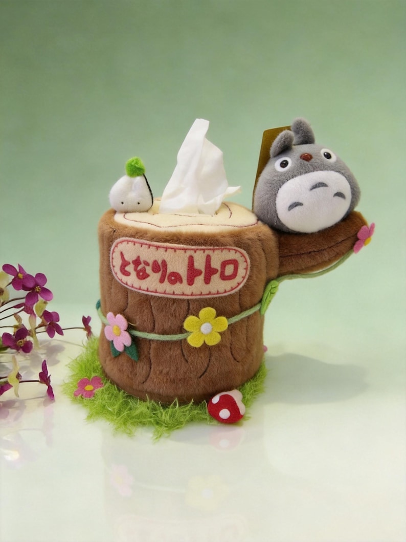 May include: A plush tissue box cover shaped like a brown tree stump with a grey Totoro character on top. The stump features pink and yellow felt flowers, a small white mushroom, and the Japanese text "となりのトトロ" embroidered on the front.