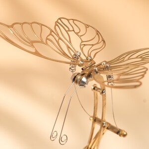 Puede incluir: Una detallada escultura de lib&eacute;lula de metal dorado con intrincados dise&ntilde;os de alas y delicadas antenas. La escultura presenta una compleja estructura mec&aacute;nica con resortes y remaches, creando una pieza art&iacute;stica &uacute;nica.