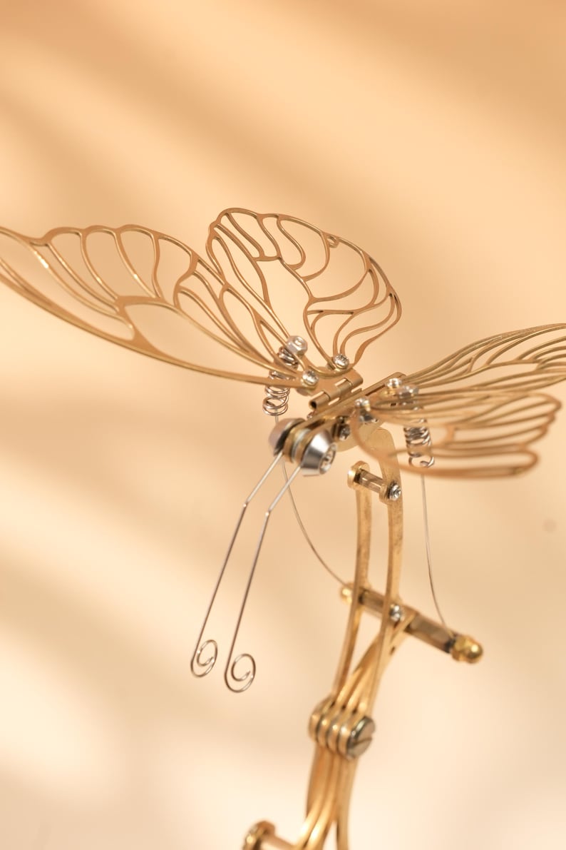 Puede incluir: Una detallada escultura de lib&eacute;lula de metal dorado con intrincados dise&ntilde;os de alas y delicadas antenas. La escultura presenta una compleja estructura mec&aacute;nica con resortes y remaches, creando una pieza art&iacute;stica &uacute;nica.