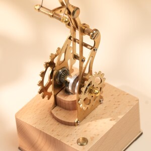 Puede incluir: Un modelo mec&aacute;nico de color lat&oacute;n con engranajes y un brazo curvo, montado sobre una base de madera clara. Los engranajes y el brazo est&aacute;n conectados por un fino alambre. El modelo es un objeto decorativo.