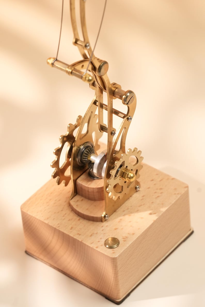 Puede incluir: Un modelo mec&aacute;nico de color lat&oacute;n con engranajes y un brazo curvo, montado sobre una base de madera clara. Los engranajes y el brazo est&aacute;n conectados por un fino alambre. El modelo es un objeto decorativo.