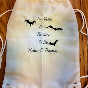 May include: White drawstring bag with black text and bat illustrations. The text reads "Too Weird To Live, Too Rare To Die, Hunter S. Thompson." The bag has white drawstrings and metal eyelets.