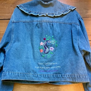 May include: A light blue denim jacket with a ruffled collar. The back features an embroidered unicorn design with flowers and the text "Now I will believe that there are unicorns."