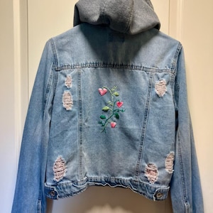 May include: A light blue denim jacket featuring a gray jersey hood, long sleeves, and a button-up front. The back displays a central embroidered vine with pink heart-shaped leaves and green stems, accented by distressed, frayed patches throughout.