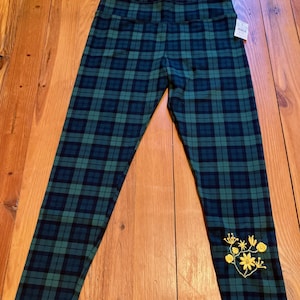 May include: Leggings featuring a dark green and navy blue plaid pattern. The garment has a high waistband and a small white retail tag attached near the top. A yellow embroidered floral design is visible on the lower left leg area.