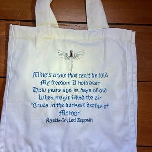 May include: Cream-colored tote bag with embroidered design and text. The design features a sword with wings above the text "Mine's a tale that can't be told...Ramble On, Led Zeppelin" in blue. The bag has two cream-colored handles.