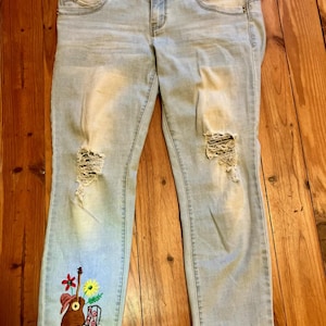 May include: Light blue denim jeans with distressed details and an embroidered design featuring a guitar, cowboy hat, flowers, and boots. The jeans have a button and zipper closure, front and back pockets.