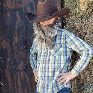 May include: A child dressed in a cowboy costume, wearing a brown cowboy hat and a long, gray beard. They are wearing a plaid shirt in blue, green, and white, along with blue jeans. The background includes a wooden wall and a hay bale.