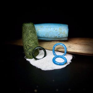 May include: An artistic composition featuring a dark green, cylindrical object, a dark green ring, and two bright blue rings. The objects rest on a bed of white sand, with a light blue, rectangular object in the background.