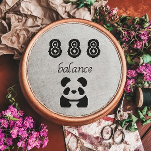 Puede incluir: Un bastidor de bordado circular de madera que contiene tela gris claro con bordado de punto de cruz negro. El diseño presenta el número 888, la palabra balance y un gráfico estilizado de un panda centrado en la superficie de la tela.