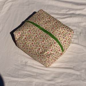 May include: A square, quilted pouch with a floral pattern of red, yellow, and pink flowers on a cream background. A bright green zipper runs across the top. The pouch is likely used for storage or travel.