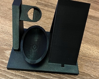 Op de afbeelding: Zwarte 3D-geprinte telefoonstandaard met een klein schaaltje en een ringhouder. De standaard heeft een gleuf voor een telefoon en een klein plankje voor een oplaadkabel.