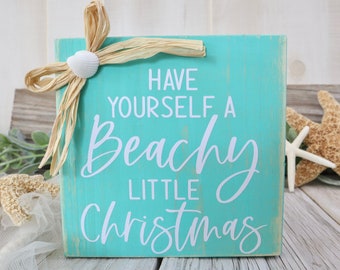 以下が含まれることがあります： 「Have Yourself a Beachy Little Christmas」の白い文字が書かれた、ターコイズ色の木製サイン。 ラフィアのリボンと貝殻で飾られています。