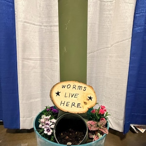 May include: A teal planter with a cylindrical green structure topped with a pink pot holding flowers. A wooden sign reading "worms live here" is attached. The planter is filled with soil and various plants.