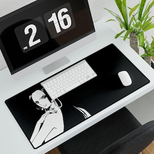 May include: Black rectangular desk mat featuring a white illustration of a woman's face. A white keyboard and mouse are on the mat. A computer monitor displays the time 2:16 PM. A green plant is in the background.