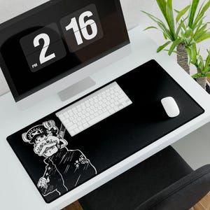May include: Black desk mat with a white cartoon character illustration. A white keyboard and mouse are on the mat. A computer monitor shows the time 2:16 PM. A green plant is in the background.