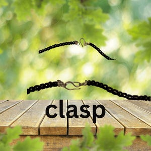 K&ouml;nnte beinhalten: Eine schwarze Perlenkette mit einem silbernen Verschluss. Das Wort "clasp" ist in schwarzen Buchstaben auf einer Holzoberfl&auml;che geschrieben.