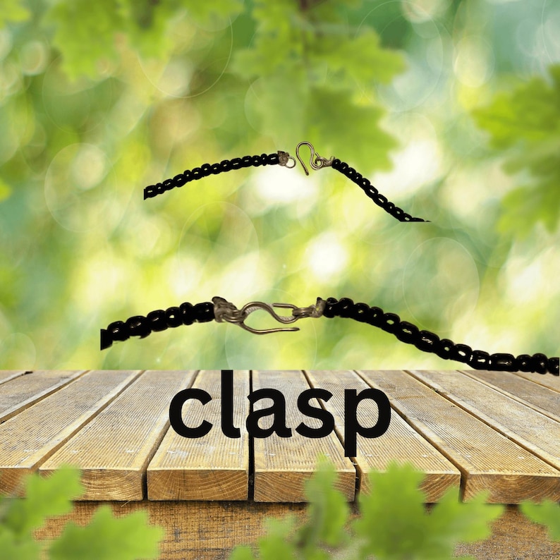 K&ouml;nnte beinhalten: Eine schwarze Perlenkette mit einem silbernen Verschluss. Das Wort "clasp" ist in schwarzen Buchstaben auf einer Holzoberfl&auml;che geschrieben.