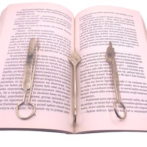 May include: Two silver letter openers rest on an open book with Polish text. The open book is on a white surface. The letter openers have decorative handles and are positioned near the text.