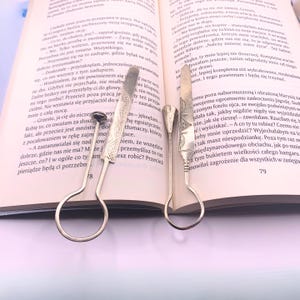 May include: Two silver-colored serving utensils with decorative handles rest on an open book. The utensils have a spoon-like end and a knife-like end. The book pages are yellowed, and the text is in Polish.