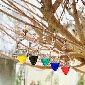 May include: Five pairs of earrings with silver metalwork and colourful, teardrop-shaped glass pendants. The pendants are yellow, black, green, blue, and red. The earrings hang from a tree branch.