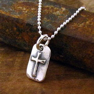 May include: A silver chain necklace with a silver pendant featuring a cross. The pendant is rectangular and has a textured surface.