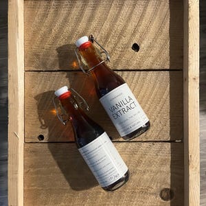 以下が含まれることがあります： 白いラベルとスイングトップクロージャーが付いた、自家製バニラエキスの小さなガラス瓶が2本。ラベルには「Homemade Vanilla Extract」と「With Love, The Taylors」と書かれています。ボトルは素朴な木製のトレイに置かれています。