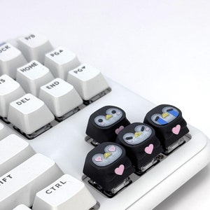 May include: Four black keycaps with penguin faces and pink hearts. The keycaps are on a white keyboard.