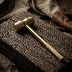 May include: A solid brass mallet featuring a cylindrical head and a slender handle with a textured, cross-hatched knurled grip. The metal surface has a polished, reflective gold-toned finish, contrasting with the dark, weathered wood grain texture.