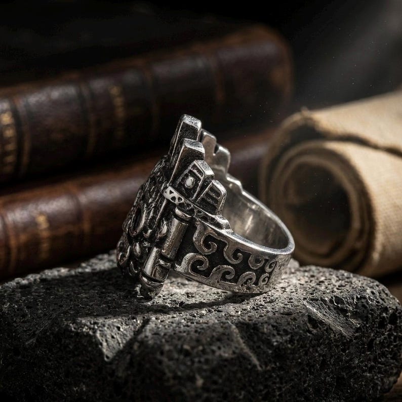 Angled view of a chunky metal Mesoamerican tribal face ring showing side band details, placed on dark volcanic stone.