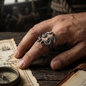 May include: A silver octopus ring with detailed tentacles encircles a finger. The ring is displayed over a map and compass, evoking a nautical or pirate theme. The ring's design is intricate and visually appealing.