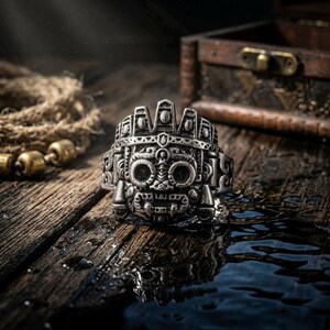 Heavy stainless steel Tláloc Aztec god of rain ring resting on wet weathered wood next to a vintage merchant chest.