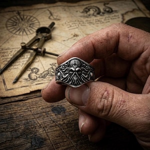 May include: A silver-colored ring with a detailed, stylized design featuring a central skull-like figure with tentacles. The ring is held in a hand against a backdrop of a vintage map and drafting compass, suggesting a nautical or fantasy theme.