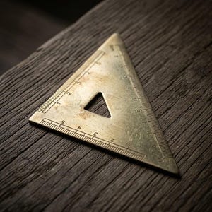 May include: A triangular brass ruler featuring etched measurement markings along its edges. The tool displays a central triangular cutout and clear numerical increments, with the longest side measuring approximately 9 centimetres in length.