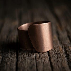 Puede incluir: Un anillo ancho de estilo envolvente hecho de cobre con un acabado mate cepillado. La banda presenta un diseño liso y redondeado que se superpone, creando una superficie sólida y continua alrededor del dedo, mostrando el tono marrón rojizo.