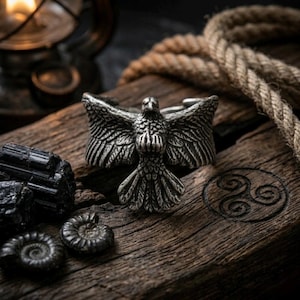 Op de afbeelding: Een zilverkleurige ring met een gedetailleerd adelaarsontwerp, met wijd gespreide vleugels. De ring ligt op een verweerd houten oppervlak, samen met zwarte kristallen, een touw en een spiraalsymbool. Het ontwerp van de ring is zeer gedetailleerd.