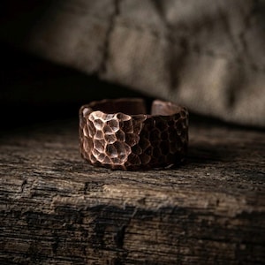 Op de afbeelding: Een brede, open ring gemaakt van koper met een uitgesproken gehamerde textuur over het gehele buitenoppervlak. Het metaal heeft een diepe, roodbruine metallic afwerking met zichtbare inkepingen die door het hameren zijn ontstaan.