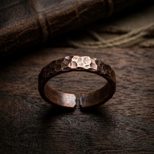 Puede incluir: Un anillo de cobre con una superficie martillada y texturizada y un diseño abierto. El metal tiene un acabado oscuro y oxidado con hendiduras visibles a lo largo de la banda, creando una apariencia rugosa. El anillo es circular con una abertura.