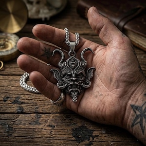 Op de afbeelding: Zilverkleurige hangerketting met een gedetailleerd, gestileerd gezichtsontwerp. De hanger hangt aan een bijpassende ketting. De ketting wordt in een hand gehouden, waarbij de ingewikkelde details van de sieraden worden benadrukt.