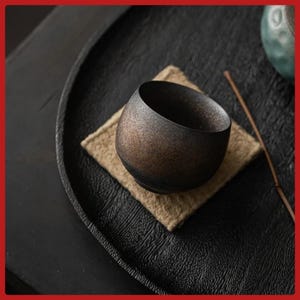 Peut inclure: Une petite tasse à thé en céramique brun foncé avec une surface texturée repose sur un dessous de verre carré beige. La tasse est placée sur un plateau noir de forme ovale. Un bâton en bois repose sur le plateau.