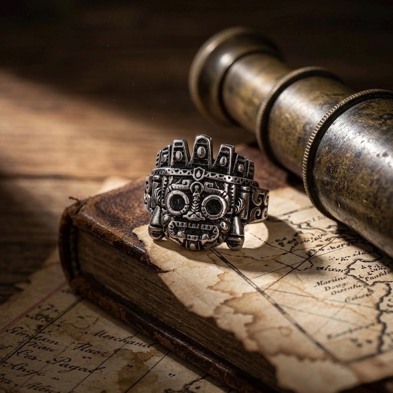 Antiqued silver Tláloc jewelry artifact resting on a vintage explorer's map alongside a leather book and brass spyglass.