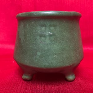 May include: A green ceramic planter with three legs. The planter has a slightly textured surface and a faded design on the front.