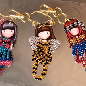 May include: Three colourful beaded keychains featuring cartoon girls with different outfits and hairstyles. Each keychain has a gold metal ring and chain.