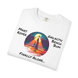 May include: White t-shirt with a circular graphic featuring a lighthouse with a beam of light, set against a colorful sunset. Text includes "Mars Rover," "Galactic Beach Bum," and "Finally Alone..."