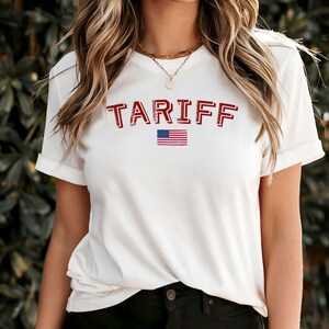 May include: White t-shirt with the word "TARIFF" in distressed red lettering and a small American flag graphic. The shirt has rolled-up sleeves and is made of a soft material.