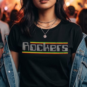 Puede incluir: Camiseta negra con la palabra "Rockers" en fuente gris desgastada, sobre una franja horizontal verde, amarilla y roja. La camiseta se usa con una chaqueta vaquera y collares superpuestos.