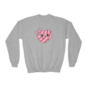 May include: A light gray sweatshirt featuring a cartoon heart-shaped concha pastry with a smiling face and a bow. The concha is pink with brown stripes. The sweatshirt has long sleeves and a crew neck.